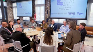 A group of people sit around two round tables pushed together in an airy, modern conference space with wood floors and columns of exposed brick between large windows. Two large screens behind the group display the Louisiana Wind Energy Week logo along with other organization logos. 
