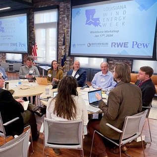 A group of people sit around two round tables pushed together in an airy, modern conference space with wood floors and columns of exposed brick between large windows. Two large screens behind the group display the Louisiana Wind Energy Week logo along with other organization logos. 