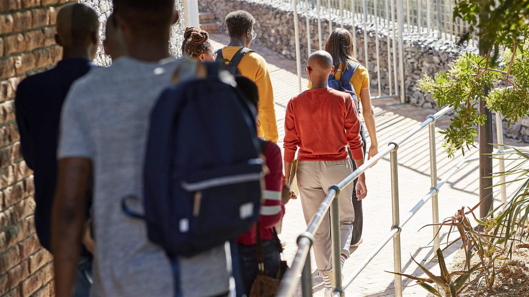 Multiethnic students wearing backpacks are shown from behind, walking on a footpath next to a brick building.