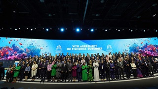 A large group of people in formal or traditional attire stands together on a stage, with a digital background depicting marine life and the large text “WTO Agreement on Fisheries Subsidies.”
