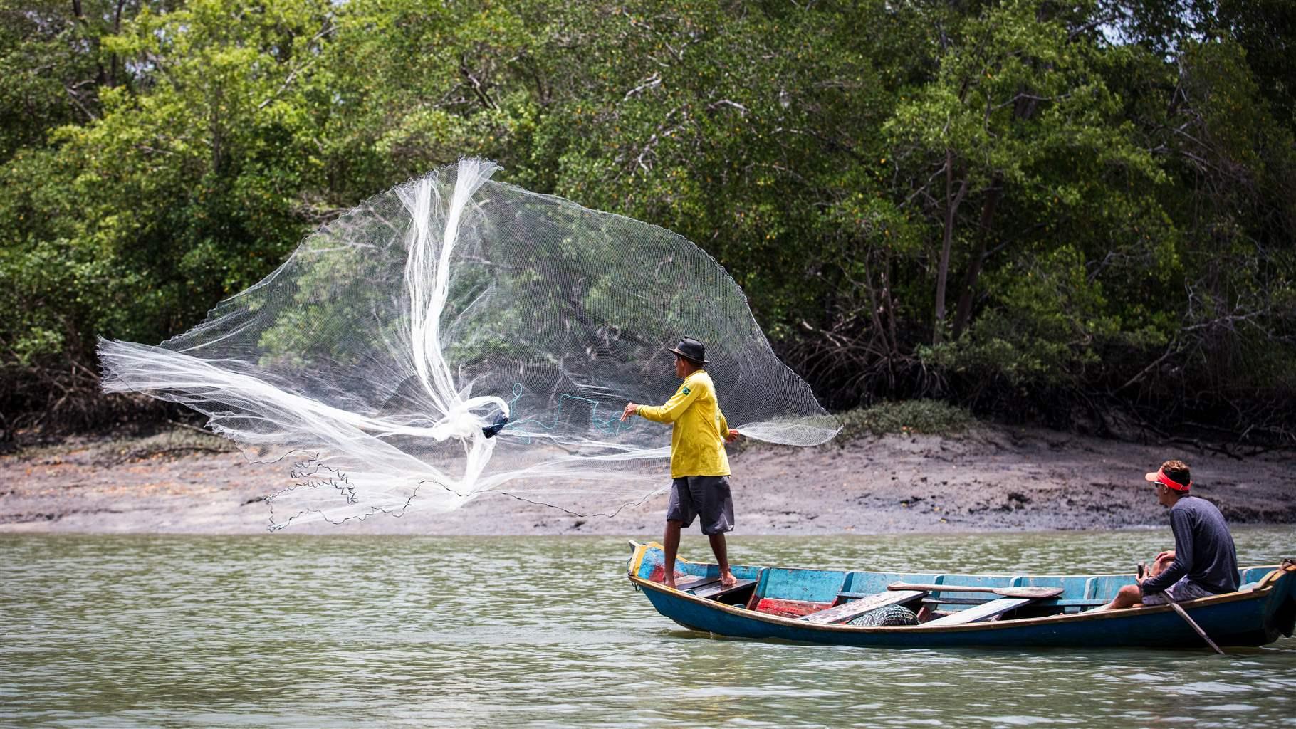 A man in shorts and a yellow shirt standing in a canoe throws a fishing net into the water while another man sits at the other end of the canoe. Onshore in the near background is a dense stand of trees.