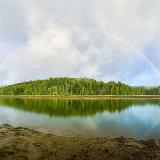 Mudflats lead to a body of water. On the horizon, a lush, forested island is crowned by the full arch of a rainbow and reflected in the still water. Low clouds hang overhead.