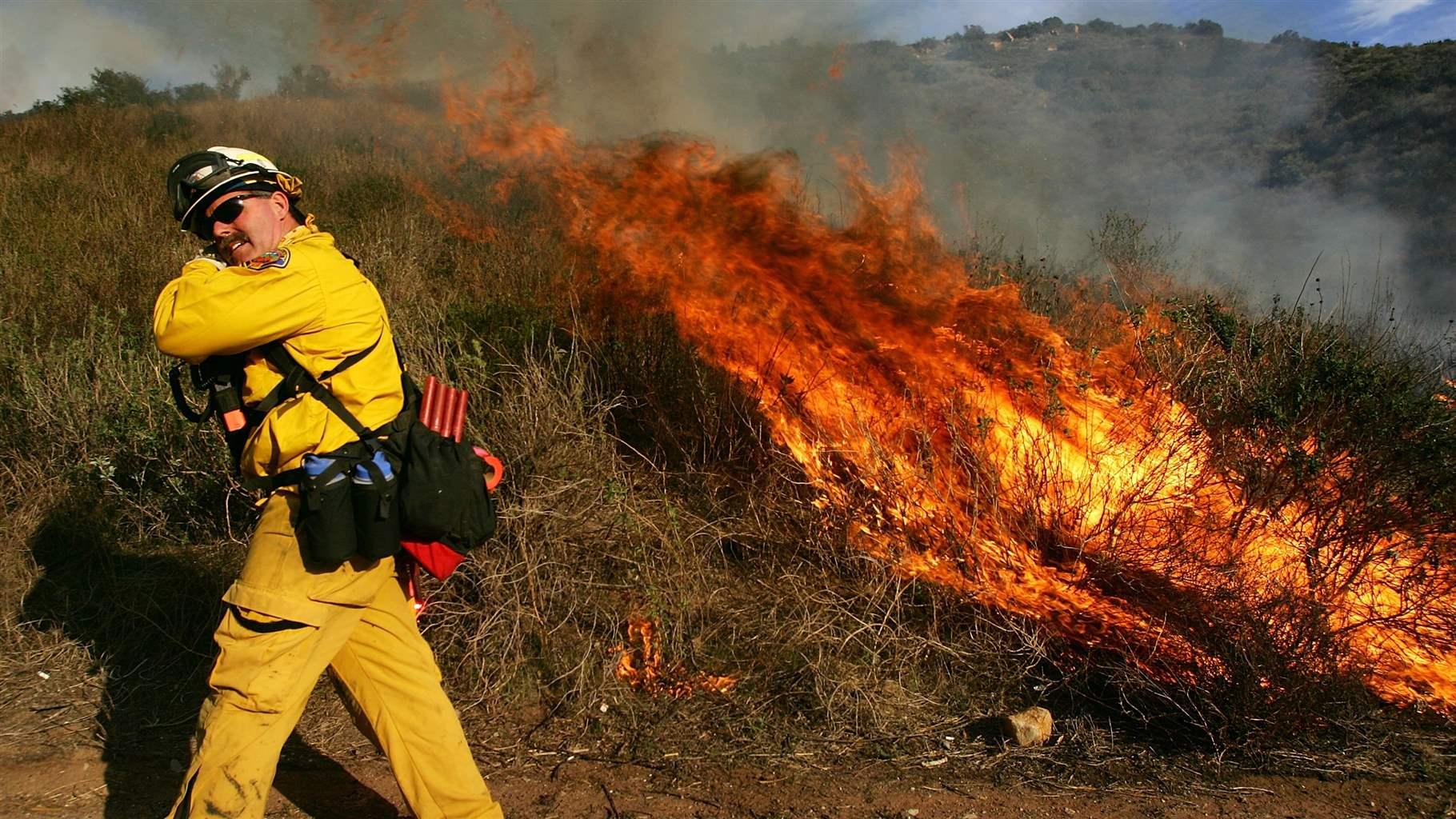 A firefighter in yellow gear turns away from a wildfire with flames and smoke visible in a grassy area.