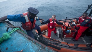An officer in an orange vest and military hat climbs down from a fishing vessel onto a dinghy. Two other officers are on the smaller boat. One reaches to help the first officer get onto the boat, and the other sits at the wheel.