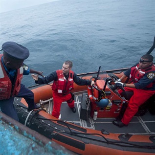 An officer in an orange vest and military hat climbs down from a fishing vessel onto a dinghy. Two other officers are on the smaller boat. One reaches to help the first officer get onto the boat, and the other sits at the wheel.