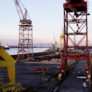 A shipyard with towers in the foreground, a barge on a river, and a blue sky in the background."