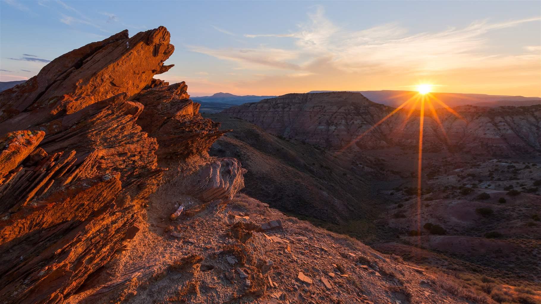 The sun starts to disappear below a hilltop, its setting rays still illuminating a craggy landscape of hills, valleys, and red rocks.