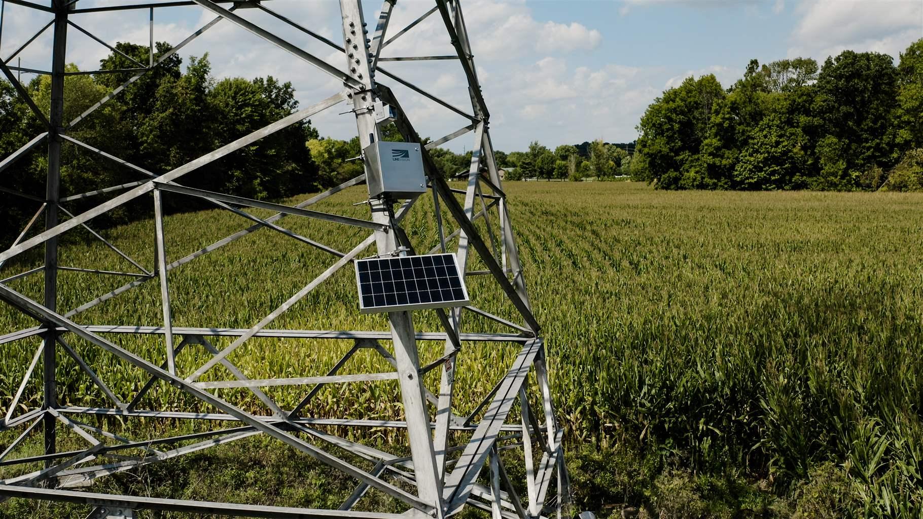 The base of a metal transmission tower stands in a green field surrounded by tall trees. A small panel and a metal box are mounted on the tower’s front-most leg. Puffy clouds punctuate a light blue sky in the background.