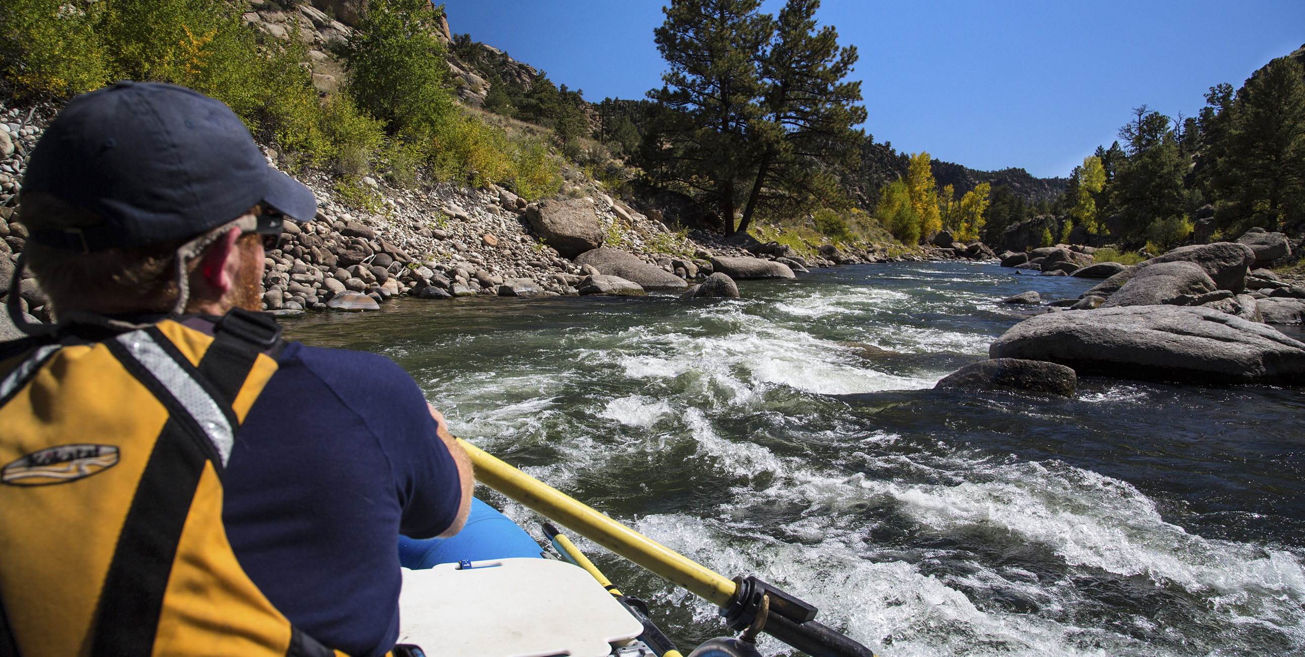 A kayaker navigates strong currents and large boulders while traveling down a narrow river surrounded by forests and clear blue sky.