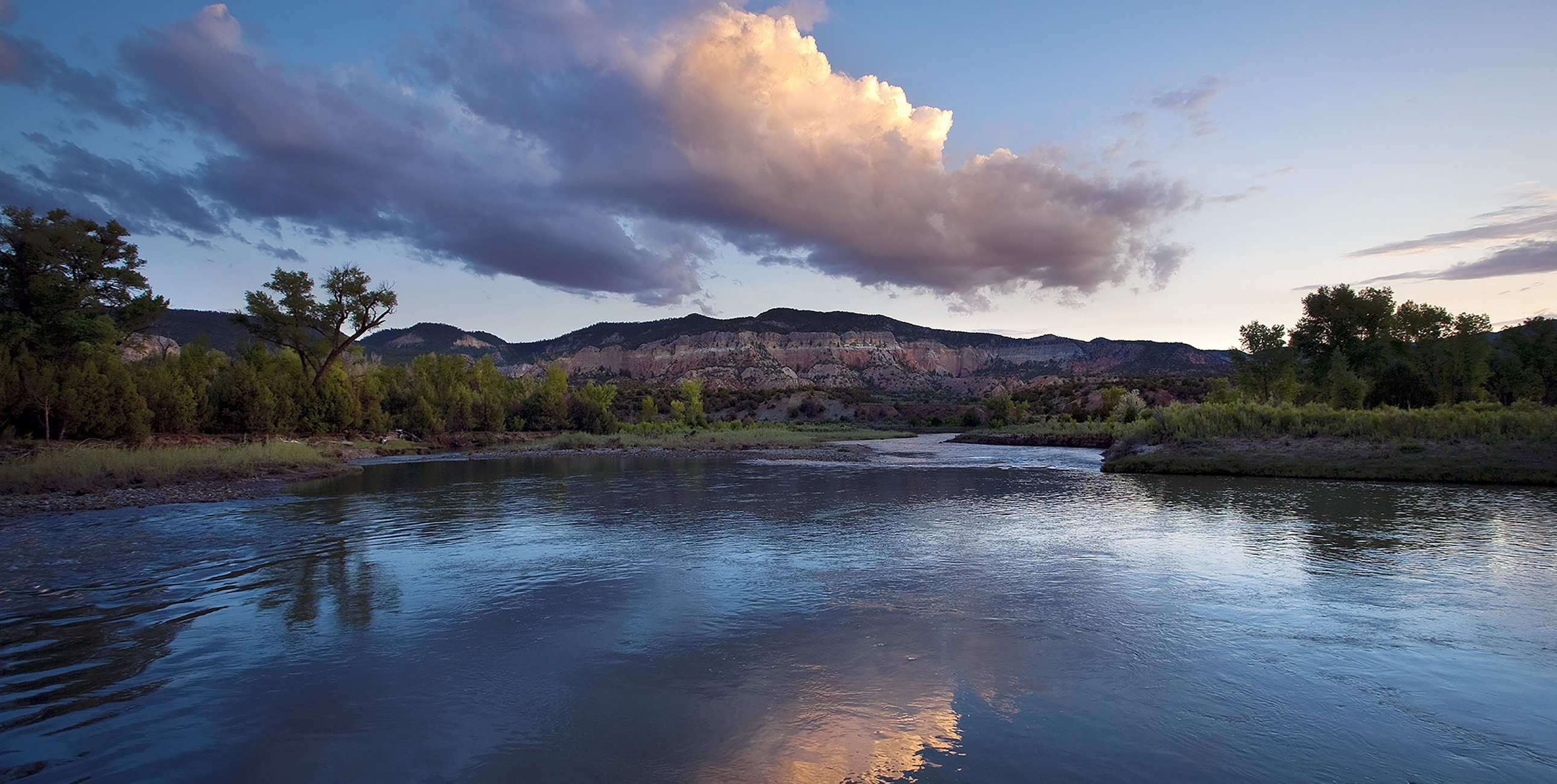 A body of water surrounded by trees, with a mountain in the background. The calm waters reflect the clouds overhead.