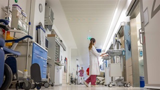 A person in a lab coat and red pants looks back at other staff members in a hospital hallway lined with medical equipment.