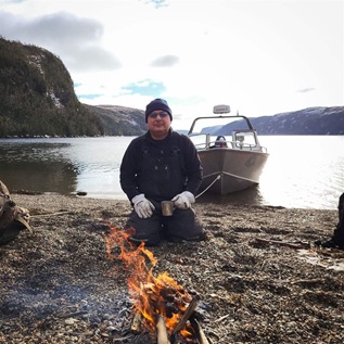 A man in a winter hat and sunglasses kneels on a rocky shoreline in front of a campfire, holding a metal mug with gloved hands. Behind him, in the water, a motorboat is moored to the shore. Cliffs rise on both sides of the water.